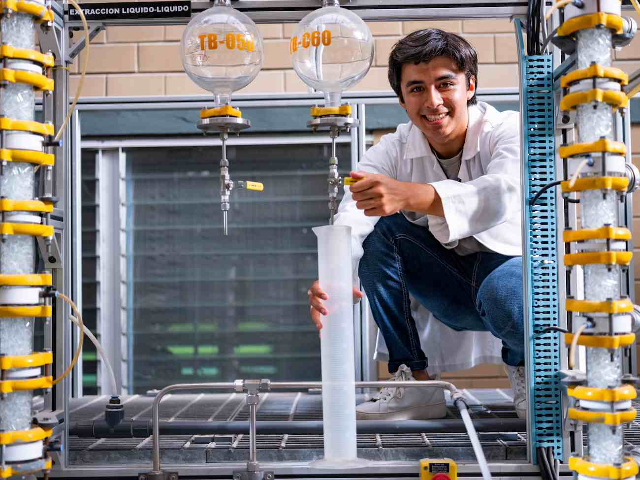 Estudiante con bata blanca acciona válvula en sistema de extracción líquido-líquido mientras llena probeta graduada en laboratorio de ingeniería química.